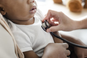 A baby, dressed in a white onesie, has their heart listened to with a stethoscope.