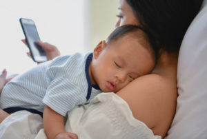 A sleeping baby nestled on his mother's shoulder as she lies down using her smartphone.