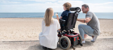 A mother and father surround their child in a wheelchair at the beach.