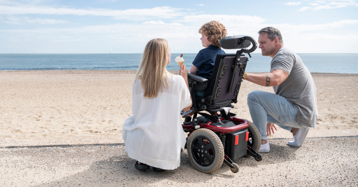 A mother and father surround their child in a wheelchair at the beach.