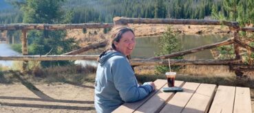 Amie Kroessig sits at the foot of the Rocky Mtns. at a wooden picnic bench with a cold brew.