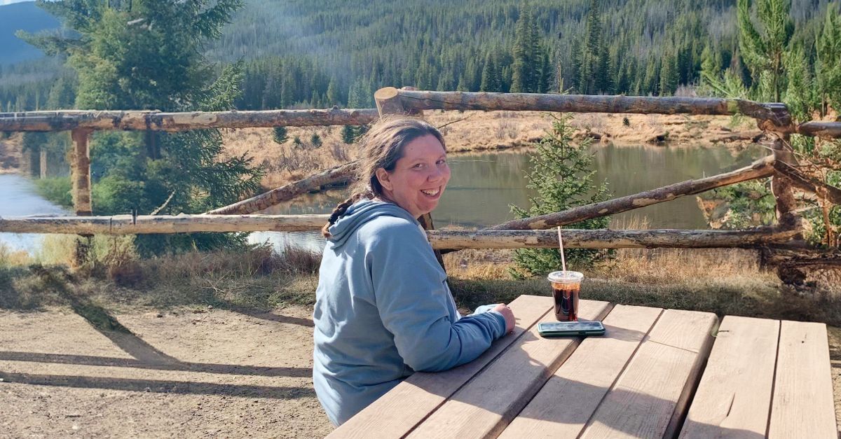 Amie Kroessig sits at the foot of the Rocky Mtns. at a wooden picnic bench with a cold brew.