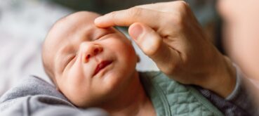 A newborn baby faces the camera with its eyes closed as an adult caresses it's forehead.