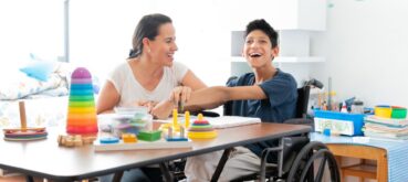 A woman who may be the child's mother or teacher, sits in a classroom, working with a wheelchair-bound child who presumably has cerebral palsy. Both are smiling.