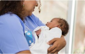 A medical professional holds a newborn baby gently, with the baby looking up at them.